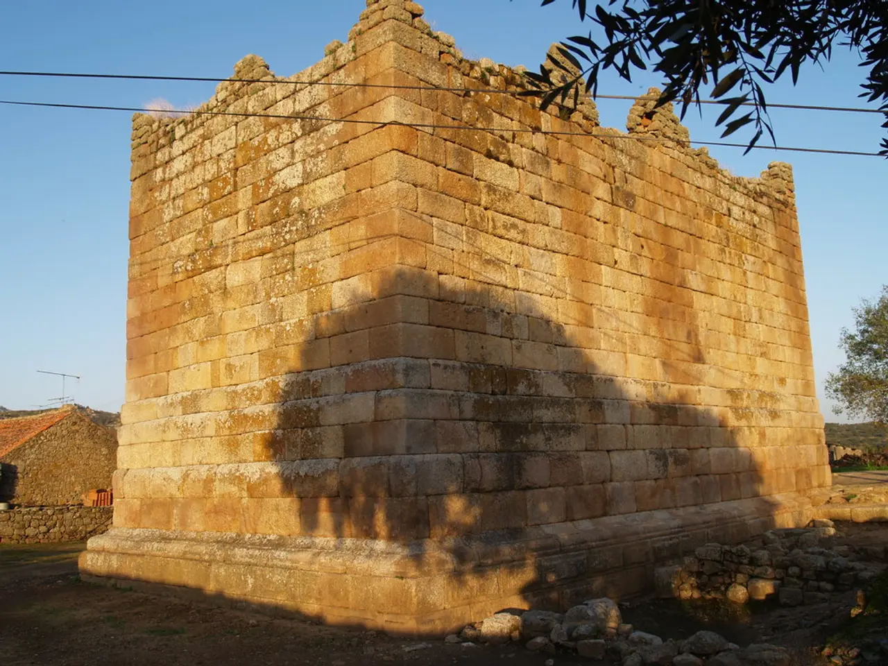 In this image we can see sky with clouds, trees, cables, for, buildings and stones on ground.
