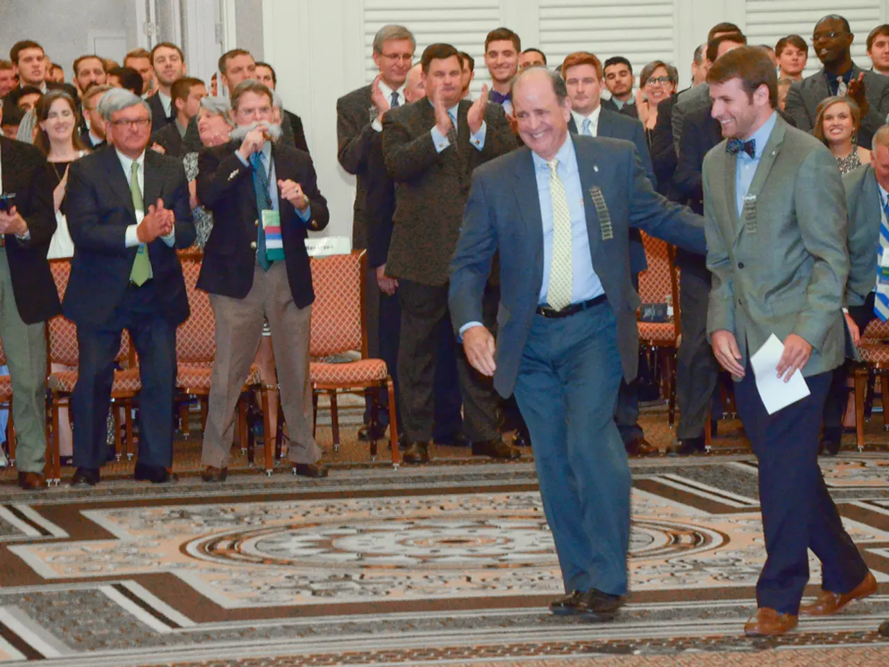 In the foreground of the picture there are two men in suits. On the right there is a microphone. In...