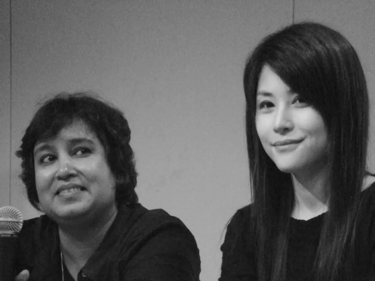 In the image there are two woman sitting on chair holding a microphone in front of a table, on...