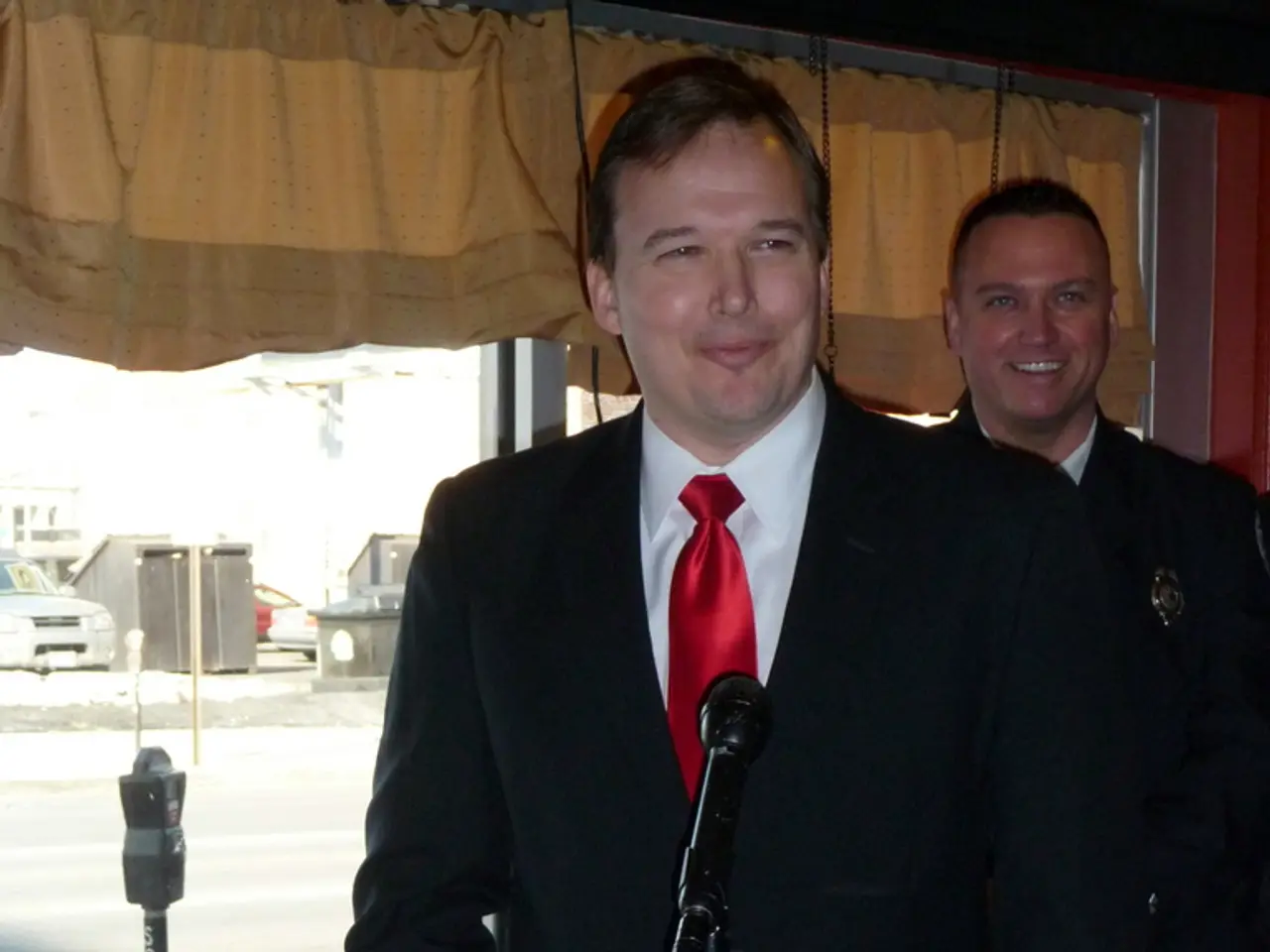 In the middle of this image, there is a person in a suit, smiling and standing in front of a table,...