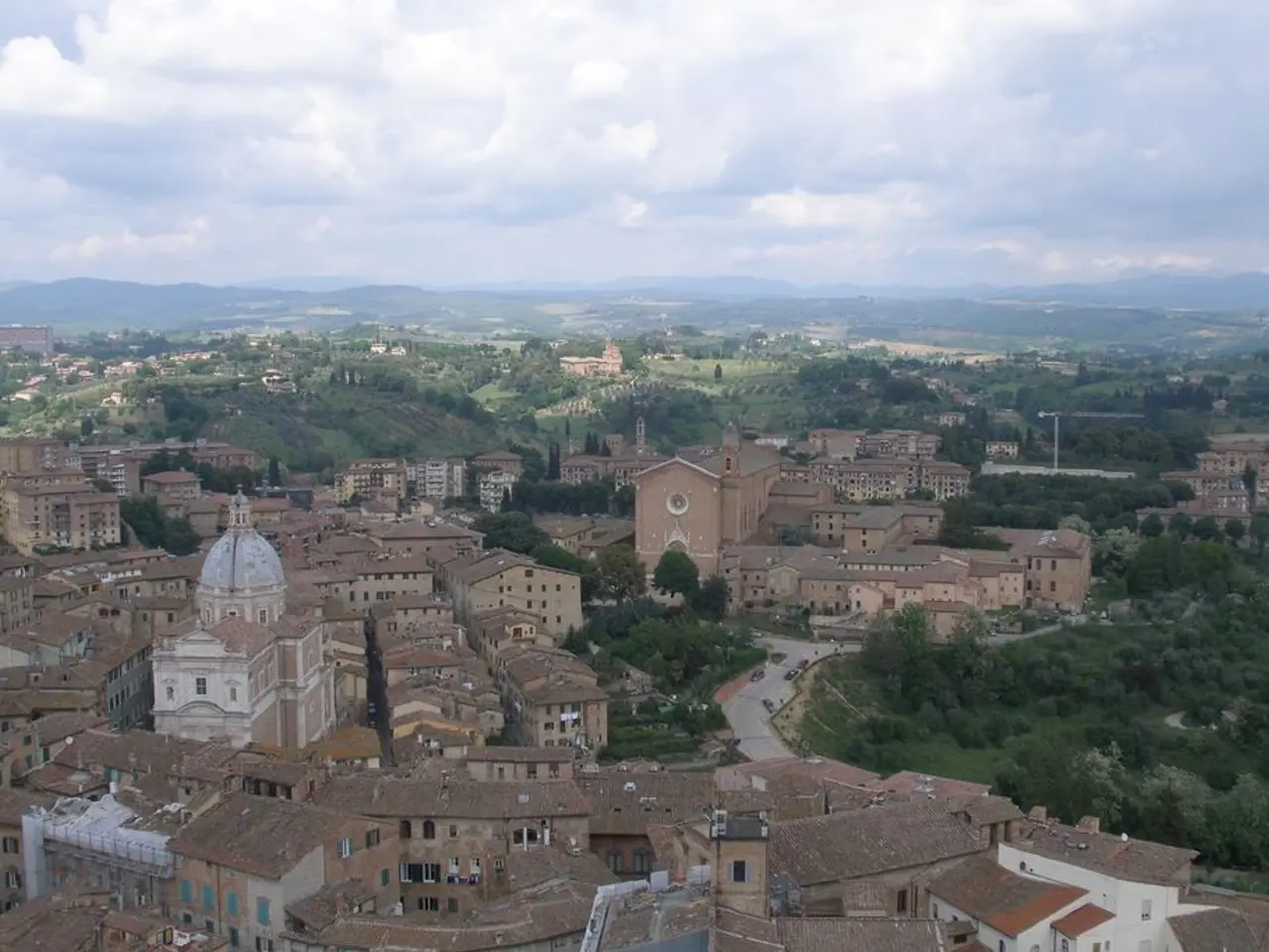 In this image there are so many buildings, trees and mountains at the back also there are clouds in...