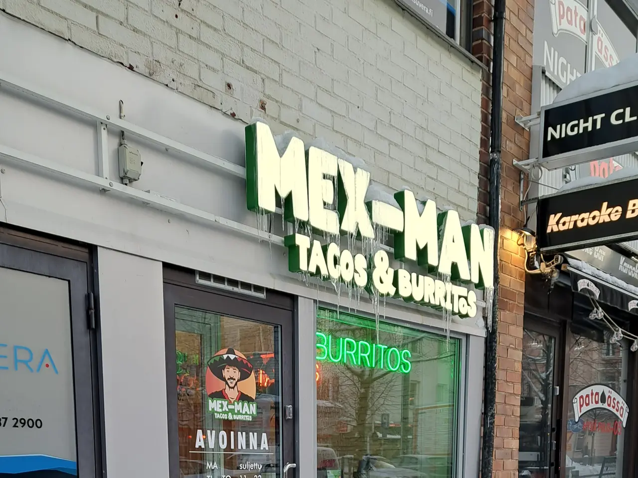 The image shows a building with glass walls and doors, boards with text, a pot with a plant, and...