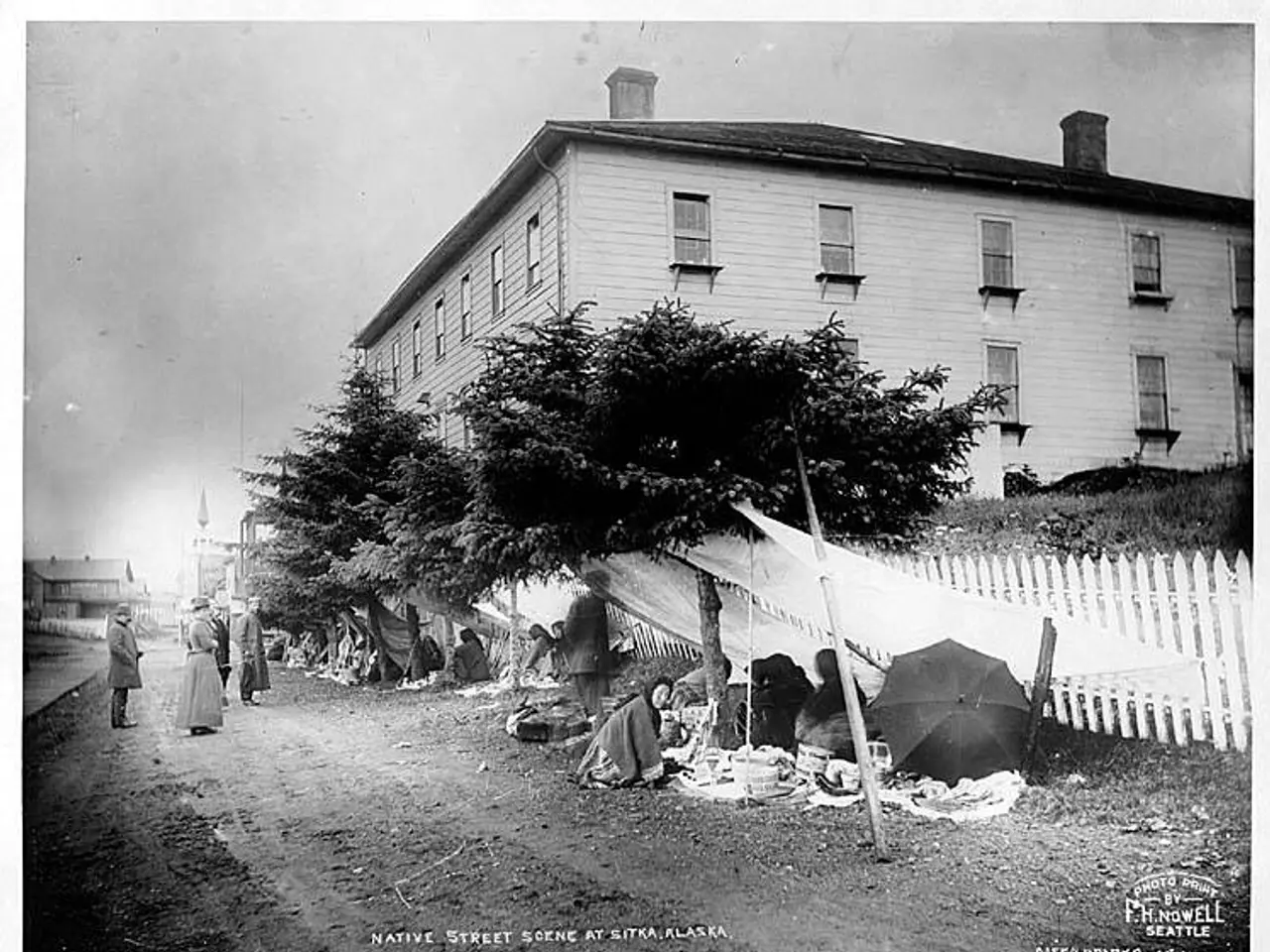 The image shows a group of people sitting on the ground in front of a white picket fence,...
