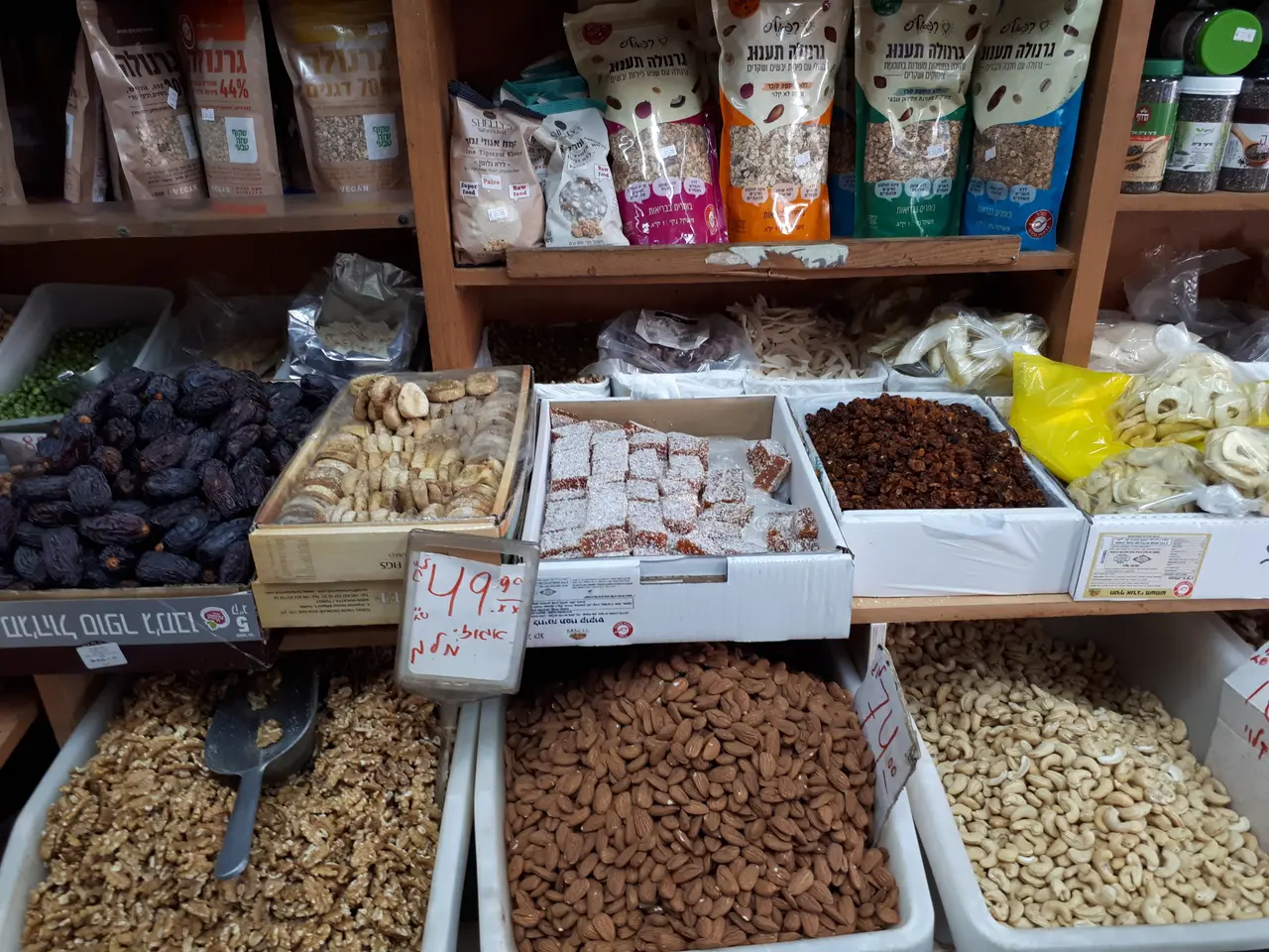 The image shows a market stall filled with a variety of nuts and dried fruits in boxes, with price...