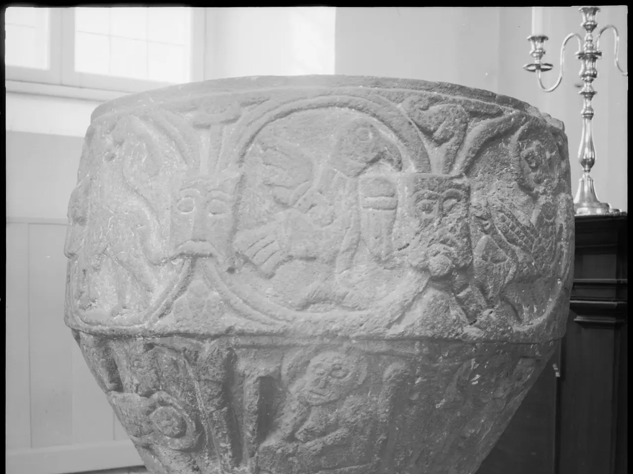 The image shows a large stone baptismal font with carvings on it, sitting atop a pedestal in front...