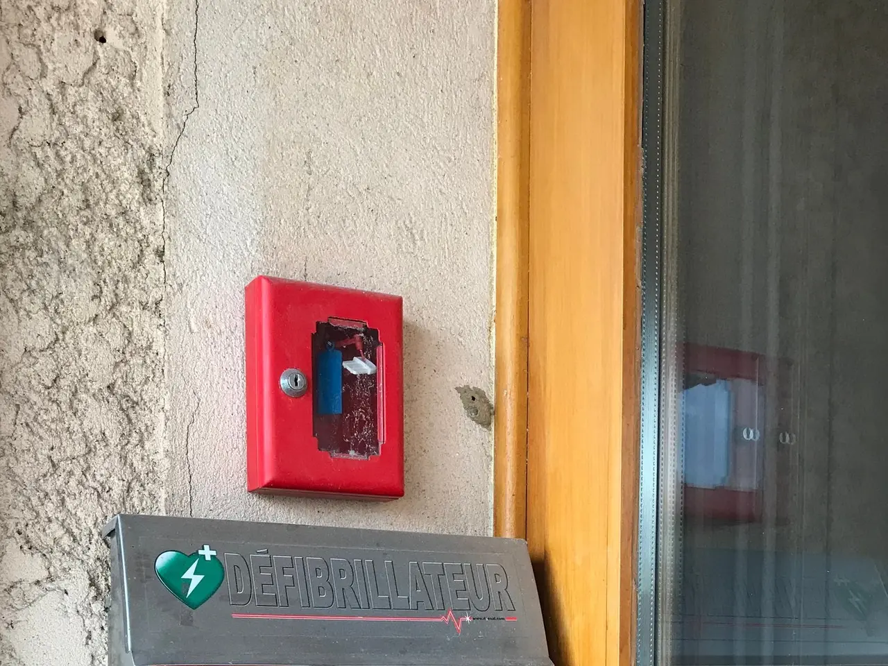 The image shows a red fire alarm mounted on the side of a building, with a glass door on the right...