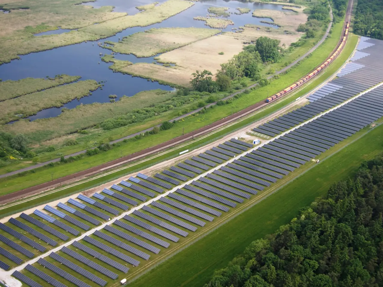 The image shows an aerial view of a large solar farm in the middle of a field, surrounded by trees,...