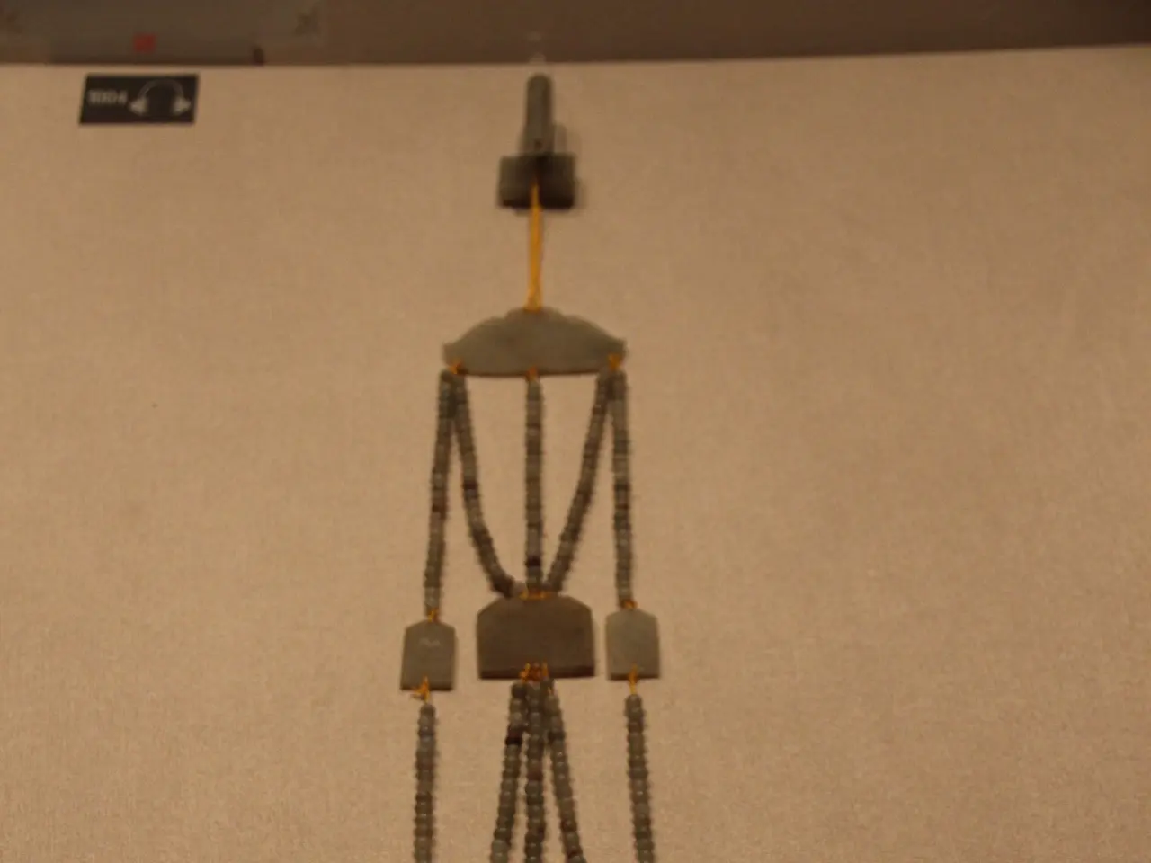 The image shows a necklace on display in a museum, with a chain of beads hanging from the wall. The...