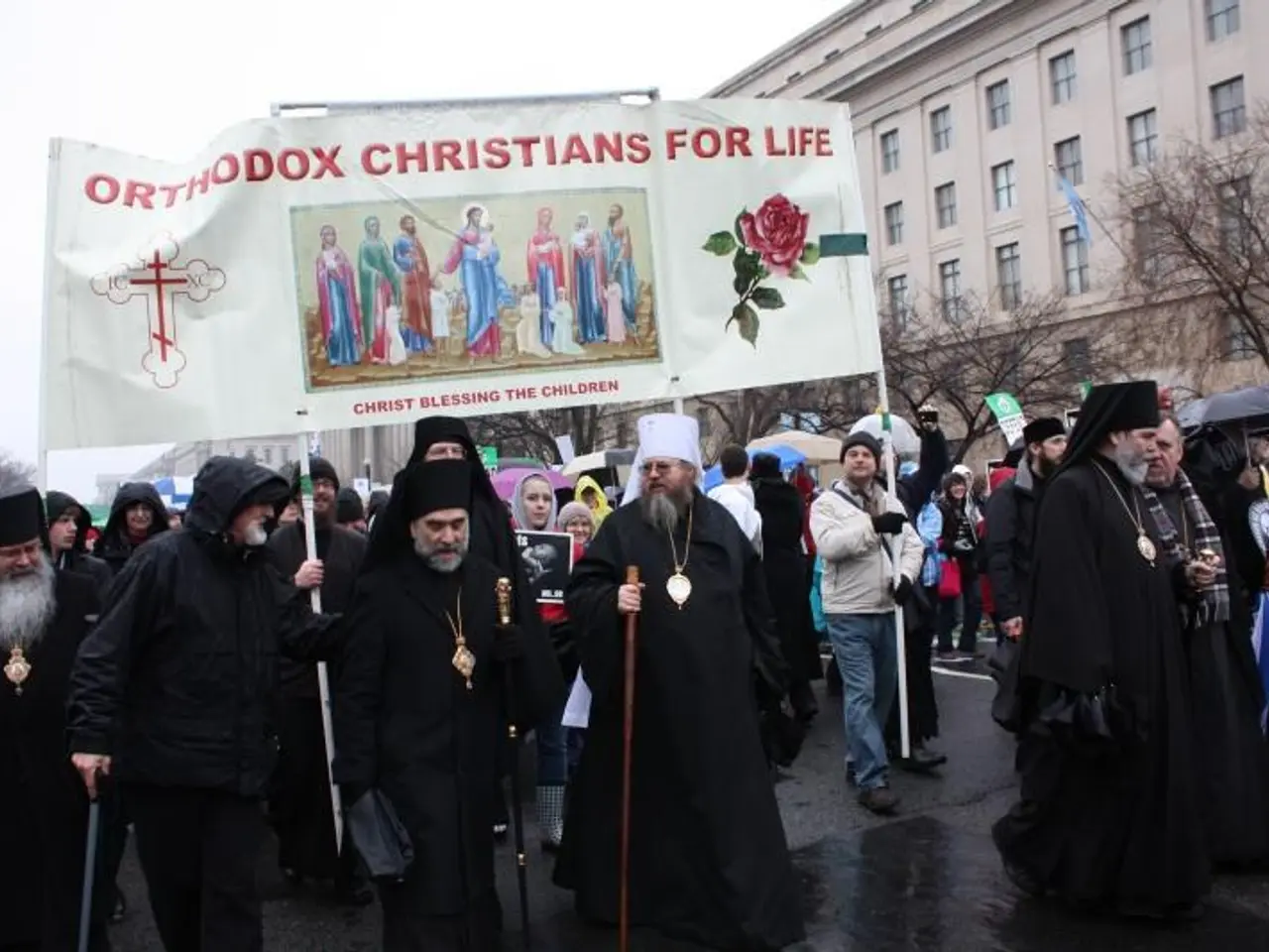 The image shows a group of orthodox Christians marching down a street, holding a banner with text...