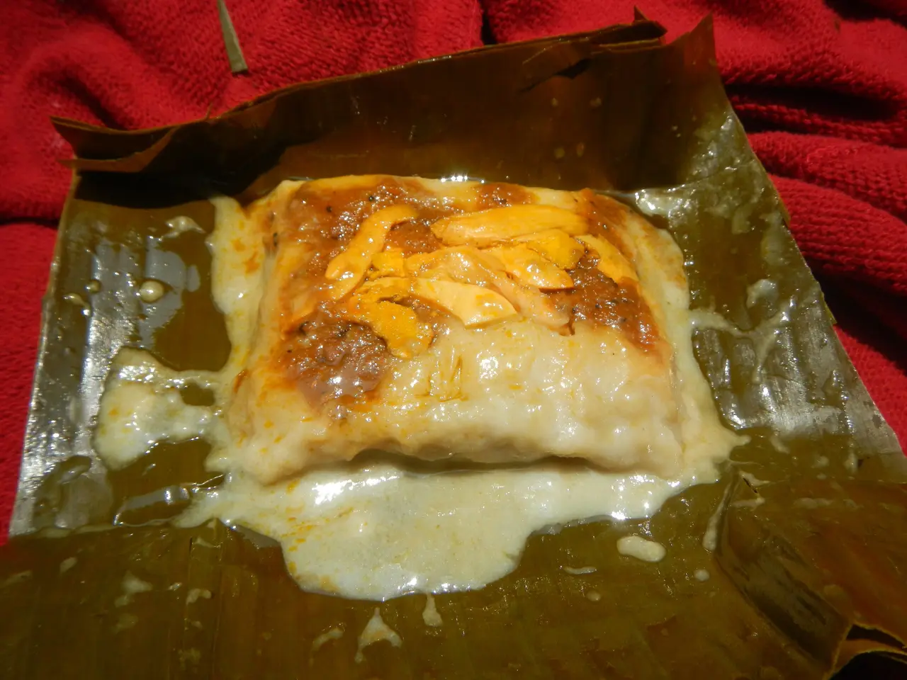 The image shows a close up of a piece of food on a banana leaf, which is placed on a red cloth. The...