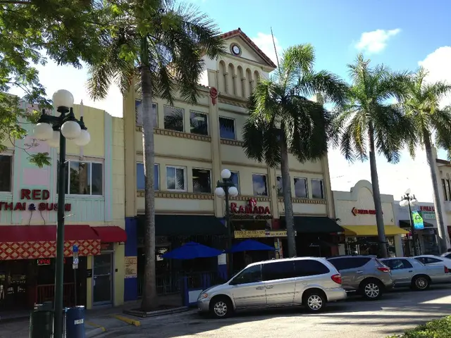 This picture shows few buildings and trees and we see couple of pole lights and cars parked on the...