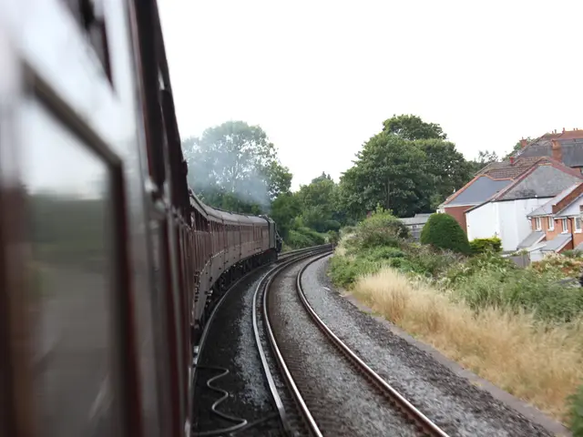 In this image we can see a train on the railway track, there are few plants and buildings on the...