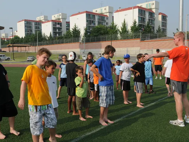 Land is covered with grass. Here we can see people. Background there are buildings, light poles,...