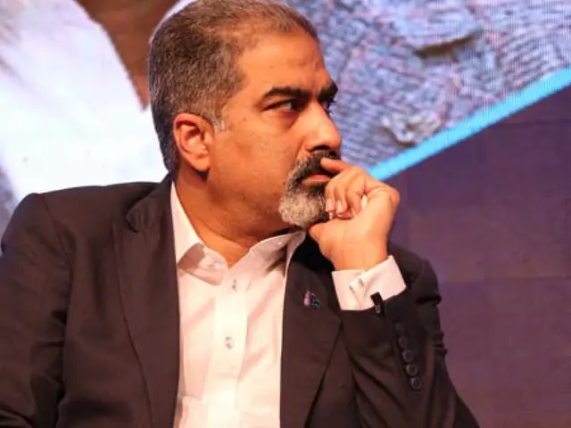 The image shows a man in a suit and tie sitting on a stage in front of a screen. He appears to be...