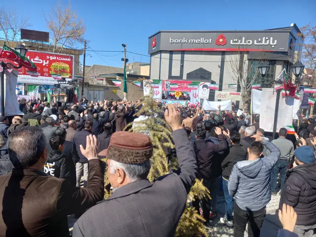 The image shows a large crowd of people standing in front of a building, holding banners and flags,...