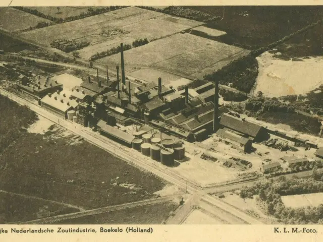 The image shows an aerial view of a factory in the middle of a field, surrounded by buildings,...