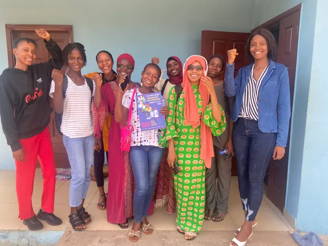 The image shows a group of women standing next to each other in front of a building, smiling and...