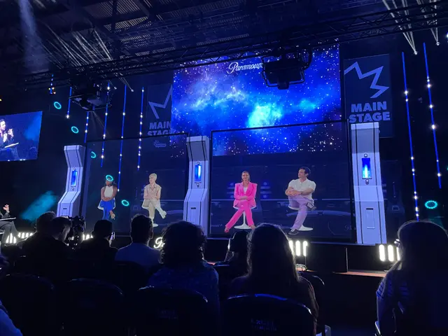 The image shows a group of people sitting in chairs in front of a large screen, illuminated by...