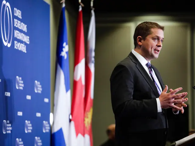 The image shows a man in a suit and tie standing at a podium with a microphone in front of him,...