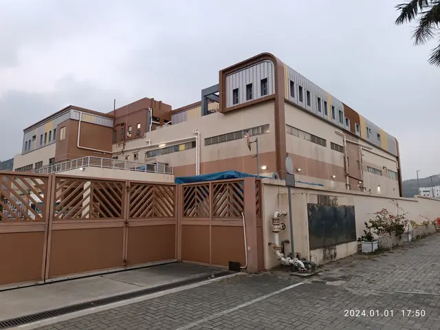 The image shows a large building with a lot of windows and a fence in front of it, surrounded by...