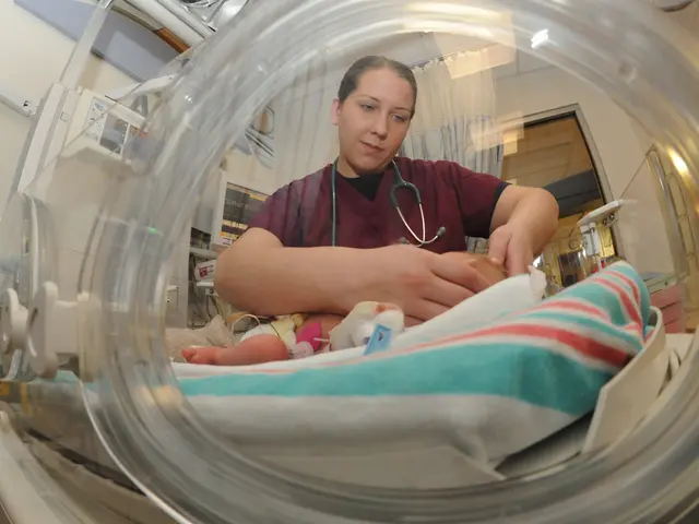 The image shows a woman in a hospital room holding a baby in an incubator. She is wearing a...