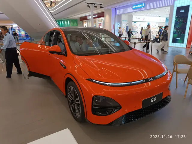 The image shows an orange car on display in a showroom, surrounded by a group of people standing on...