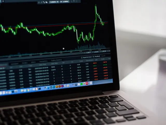 The image shows a laptop computer sitting on top of a white desk, with a stock market graph...
