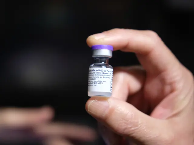 The image shows a person holding a vial of Covid-19 vaccine in their hand, with a blurred...