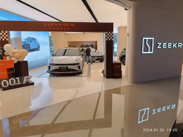 The image shows a car showroom with a car on display in the center, surrounded by people standing...