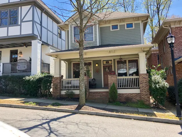 The image shows a two story house with a car parked in front of it, surrounded by trees, plants,...