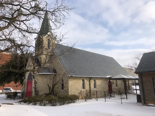 The image shows a small church with a steeple in the middle of a snowy street, surrounded by houses...