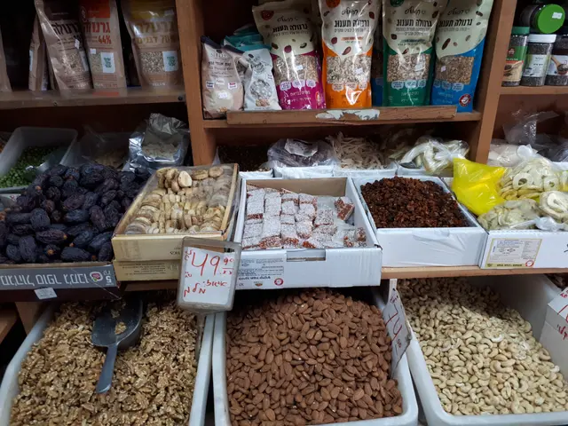 The image shows a market stall filled with a variety of nuts and dried fruits in boxes, with price...
