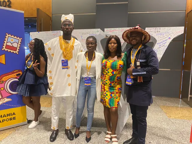 The image shows a group of people standing next to each other in front of a sign, wearing tags and...