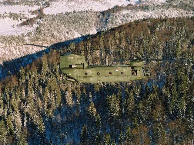 The image shows a military helicopter flying over a forest filled with trees and snow-covered...
