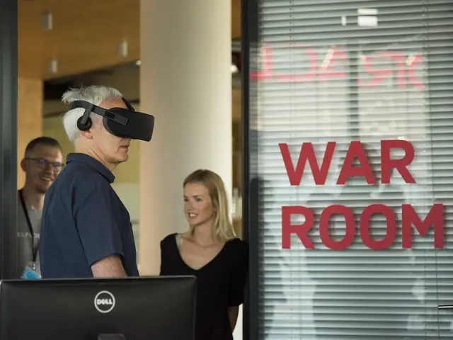 The image shows a man wearing a VR headset standing in front of a computer in a war room. He is...