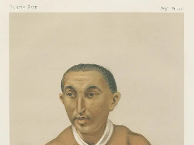 The image shows a man sitting at a table with his hands clasped in front of him. He is wearing a...