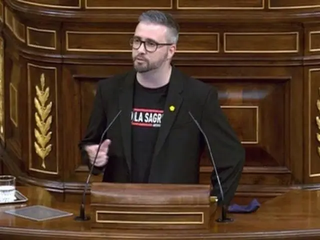 The image shows a man standing at a podium in front of a wooden wall, wearing a black blazer and...