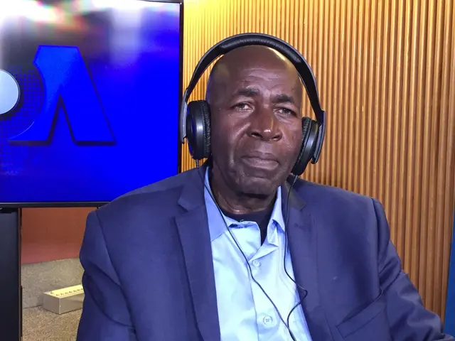 The image shows a man in a suit and headphones sitting in front of a TV, with a wooden wall in the...