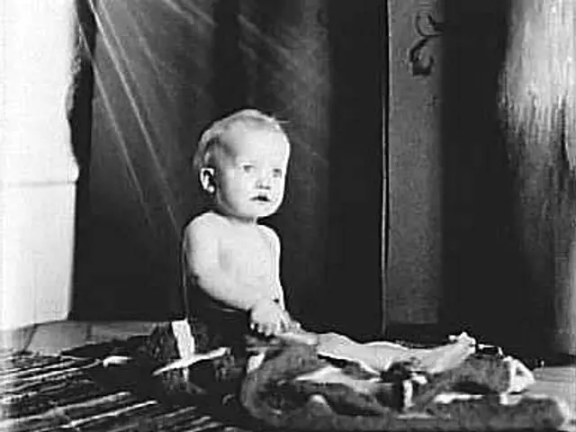 The image shows an old black and white photo of a baby sitting on the floor in front of a wall. The...