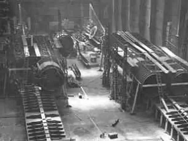 The image shows a black and white photo of a factory with a large room filled with lots of pipes,...