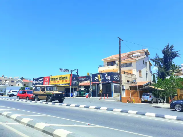 The image shows a street with cars parked on the side of it, a divider, traffic cones, poles,...