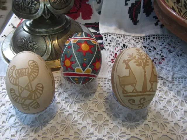 The image shows three decorated eggs sitting on top of a table covered with a cloth, with a bowl...