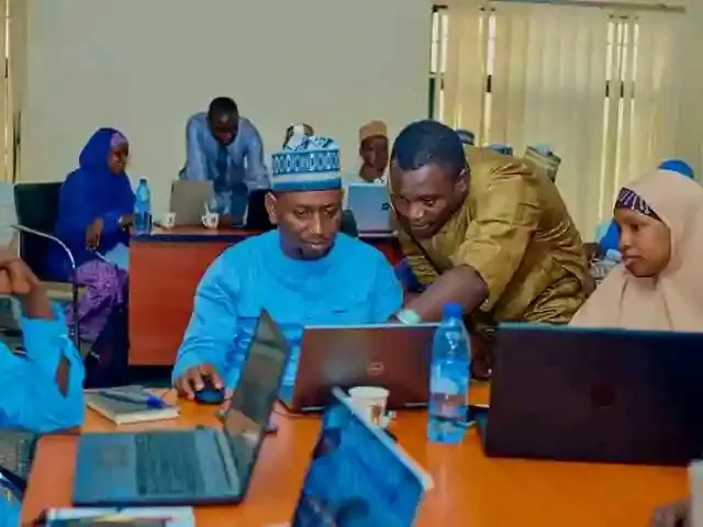 The image shows a group of people sitting around a table with laptops, bottles, cups, books, pens,...