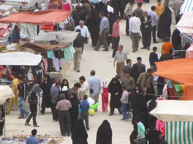 The image shows a crowd of people walking through a bustling bazaar, with stalls on either side of...