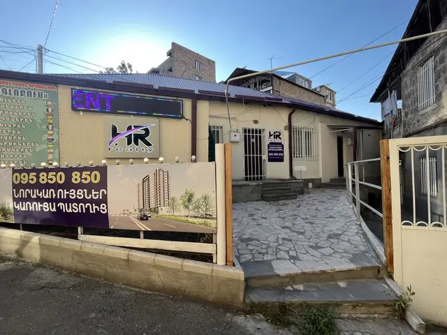The image shows a building with a sign in front of it, surrounded by a metal fence, a gate, some...