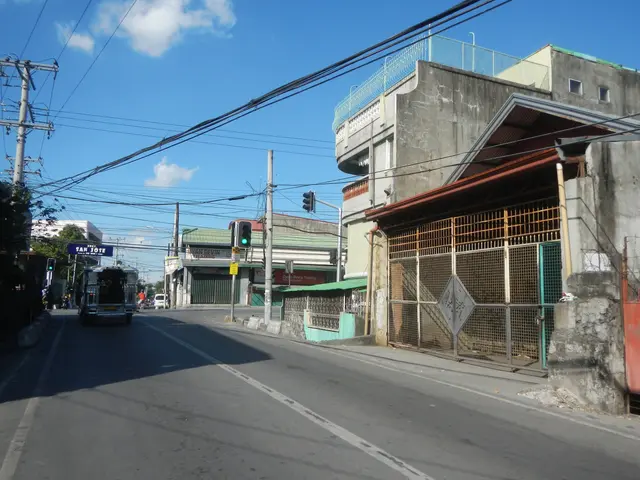 The image shows a city street with a bus driving down it, surrounded by buildings, electric poles...