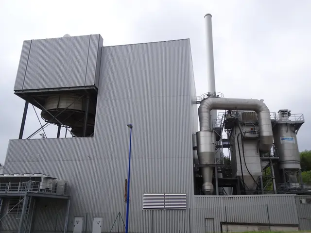 The image shows a large industrial building with a lot of pipes on top of it, surrounded by a road,...