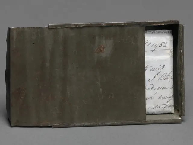 The image shows a metal box with a piece of paper inside of it on a grey surface. The paper has...