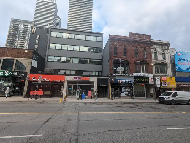 The image shows a city street with tall buildings in the background, vehicles on the road, people...