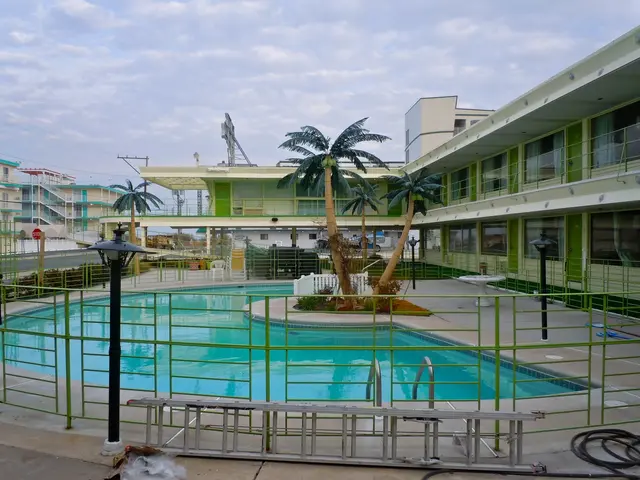 The image shows a large swimming pool in front of a building with palm trees, surrounded by a metal...